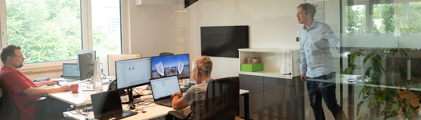 Two employees are sitting at their desks, another comes in the door