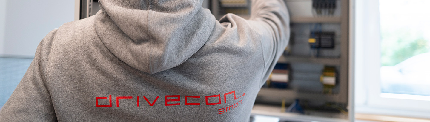 Rückenansicht eines Hoodies mit DriveCon-Logo, der Mitarbeiter steht vor einem Schaltschrank